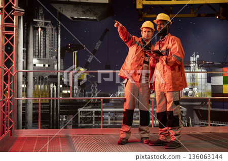 Diverse engineering experts in orange jackets and hard hats analyze operations on offshore rig, ensuring compliance with oil and gas industry standards used for fuel extraction. 136063144
