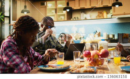 Busy african american working dad typing on laptop at table with his family, ignoring his small children and wife during breakfast. Distracted preoccupied father distant with his two kids. Camera A. 136063158