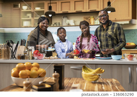 Portrait of adorable black family enjoying pizza day and cooking together in the kitchen. Children and parents sprinkling fresh vegetables and other ingredients to create delicious food. Portrait of adorable black family enjoying pizza day and cooking together in the kitchen. Children and parents sprinkling fresh vegetables and other ingredients to create delicious food. 136063290