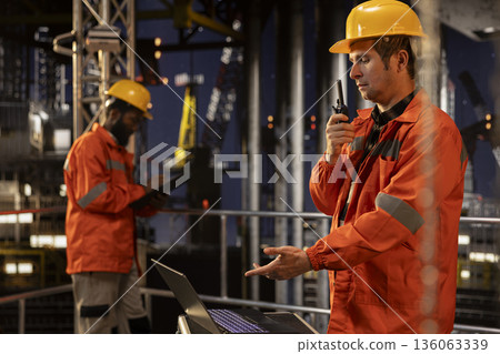 Skilled offshore engineer talking through walkie talkie at night for drilling rig systems monitoring activity and promoting petrol control and extraction practices. Oil pumping. Skilled offshore engineer talking through walkie talkie at night for drilling rig systems monitoring activity and promoting petrol control and extraction practices. Oil pumping. 136063339