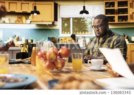 African american unfocused father paying attention to work on laptop and ignoring his family at the breakfast table. Absorbed distant working dad distracted by gadget, neglecting kids. 136063343