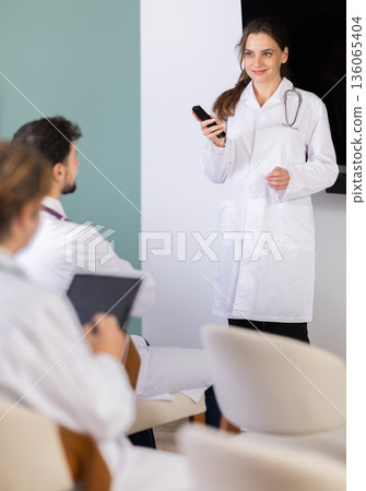 Female doctor lecturer communicates and addressed to attendees, control projector using remote control Female doctor lecturer communicates and addressed to attendees, control projector using remote control 136065404