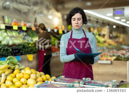 Woman store employee with folder of documents takes inventory in vegetable store Woman store employee with folder of documents takes inventory in vegetable store 136065583