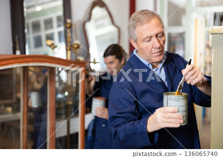 Skilled old furniture repair specialist in uniform restoring and painting cupboard furniture during workday in workshop 136065740