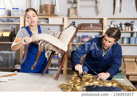 Male restorer treats wooden frame of vintage mirror with protective varnish. While woman restorer restores upholstery of vintage chair Male restorer treats wooden frame of vintage mirror with protective varnish. While woman restorer restores upholstery of vintage chair 136065857