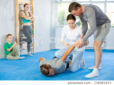 Woman is learning how to grab opponent's hand in prone position during self-defense class, in background girl and boy watch sparring 136065873