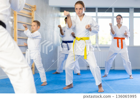 Teen girl with her family in kimonos and colored belts practicing karate with punches during group martial arts class in gym, accompanied by trainer 136065995