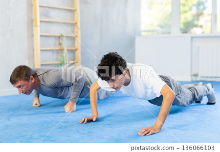 Underage boy and middle-aged trainer practicing plank pose in sports hall 136066103