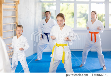 Teen girl with her family in kimonos and colored belts practicing karate with punches during group martial arts class in gym, accompanied by trainer Teen girl with her family in kimonos and colored belts practicing karate with punches during group martial arts class in gym, accompanied by trainer 136066159