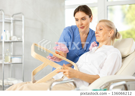 Mature woman looking at mirror while cosmetologist showing results of facial procedure Mature woman looking at mirror while cosmetologist showing results of facial procedure 136066212