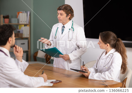 Working atmosphere, medical colleagues gathering at table, and man doctor stands up to speech 136066225