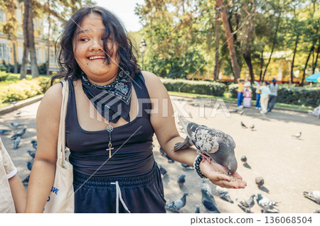 young girl with long brown hair feeds a pigeons from her hand in an urban park. The scene is lively and vibrant. young girl with long brown hair feeds a pigeons from her hand in an urban park. The scene is lively and vibrant. 136068504