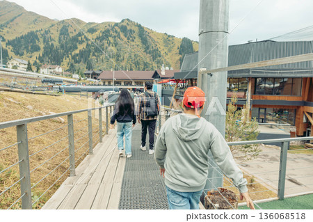family walking along wooden pathway at mountain resort, parents and child from behind exploring alpine village with cable car station and modern buildings, outdoor travel scene, concept of tourism 136068518