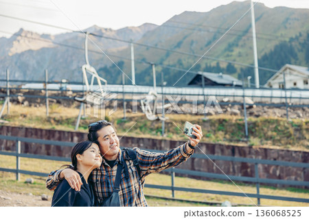 young Asian couple stands together in a mountainous area. They smile while take a selfie near a cable car station, enjoying their vacation in nature. young Asian couple stands together in a mountainous area. They smile while take a selfie near a cable car station, enjoying their vacation in nature. 136068525