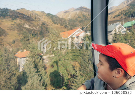 young Asian boy with short black hair wearing a red cap looks out of a cable car at mountains, houses and trees during vacation. young Asian boy with short black hair wearing a red cap looks out of a cable car at mountains, houses and trees during vacation. 136068583