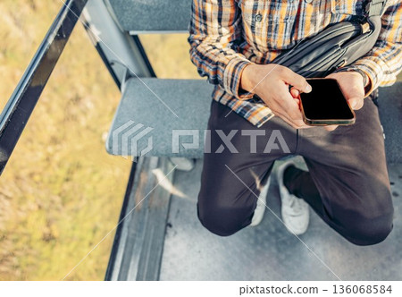 man with dark hair and a plaid shirt sits in a cable car, using a smartphone. The scene captures a vacation atmosphere in the mountains. man with dark hair and a plaid shirt sits in a cable car, using a smartphone. The scene captures a vacation atmosphere in the mountains. 136068584