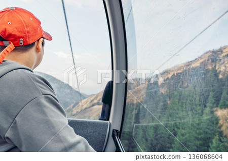 young Asian boy with short black hair wearing a red cap looks out of a cable car at mountains and trees during vacation. young Asian boy with short black hair wearing a red cap looks out of a cable car at mountains and trees during vacation. 136068604