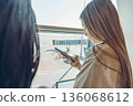 Two young women are waiting at an airport. They are looking out of large window. One has long brown hair, and the other has long black hair. 136068612
