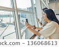 Two young women are waiting at an airport. They are looking out of large window. One has long brown hair, and the other has long black hair. Both are focused on the view 136068613