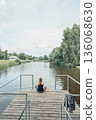 woman sitting cross-legged on wooden dock over calm river, wearing sportswear, back view, summer greenery, countryside houses, meditation, morning ritual, nature therapy, vertical 136068630
