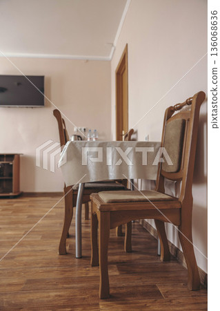 A cozy hotel dining area with a wooden table covered in a white tablecloth. Two wooden chairs are positioned around the table. A television is mounted on the wall. 136068636