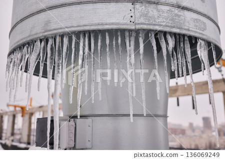 Metal vent with hanging icicles, Frozen icicles draping metallic chimney vent, Closeup of icy metal vent and chimney under overcast sky in winter storm conditions 136069249