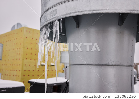 Metal vent with hanging icicles, Frozen icicles draping metallic chimney vent, Closeup of icy metal vent and chimney under overcast sky in winter storm conditions 136069250