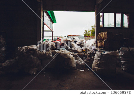 Piled plastic bags silhouetted in doorway, translucent film catching light, interior darkness framing heap, sacks and loose film spilling inward, moody sorting scene 136069578