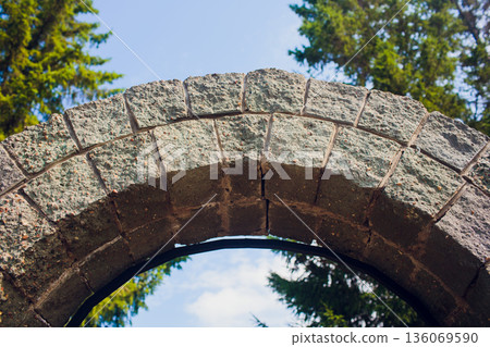 Ancient archway surrounded by lush trees and sky. Calm and peaceful outdoor archway constructed of historic stones amid flourishing foliage and expansive blue sky 136069590