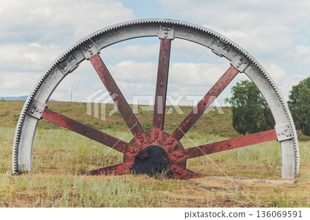 Old wheel scene. Abandoned rusty wheel surrounded by peaceful natural landscape scene. Old rusted wheel partially buried in meadow with rolling hills and cloudy sky above 136069591