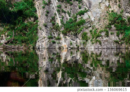 Tranquil landscape with reflective water. Quiet cliffside scene with lush plant life. Serene image of limestone precipice mirrored perfectly on undisturbed water surface 136069615