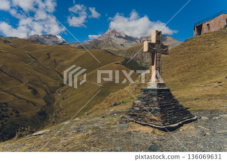 Serene stone cross on exposed mountain summit under vivid azure sky. Solitary stone monument perched on rugged ridge amidst layered peaks and clear alpine light Serene stone cross on exposed mountain summit under vivid azure sky. Solitary stone monument perched on rugged ridge amidst layered peaks and clear alpine light 136069631