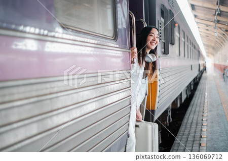 A cheerful young woman with a suitcase looks out from a train door, excited for her journey at a bustling station. A cheerful young woman with a suitcase looks out from a train door, excited for her journey at a bustling station. 136069712