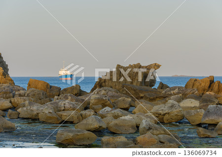 A rocky coastline and a blue ship floating offshore A rocky coastline and a blue ship floating offshore 136069734