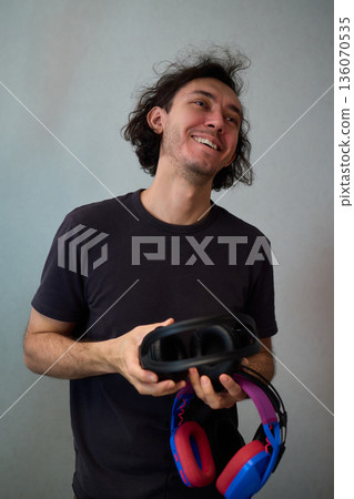 Happy guy with headphones, Casual man showcases vibrant headphones, Casually dressed man with curly hair showcases playful headphones during informal indoor photography session Happy guy with headphones, Casual man showcases vibrant headphones, Casually dressed man with curly hair showcases playful headphones during informal indoor photography session 136070535