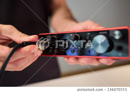 Preparation scene of connecting instrument cable to audio interface for recording, Detailed view of hand plugging instrument cable into red audio interface in studio setting 136070553