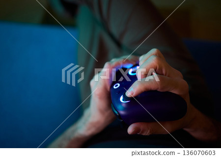 Comfortable man enjoying gameplay under colorful neon illumination, Chill indoor scene featuring smiling individual engaged in video game entertainment amid vivid purple lights Comfortable man enjoying gameplay under colorful neon illumination, Chill indoor scene featuring smiling individual engaged in video game entertainment amid vivid purple lights 136070603