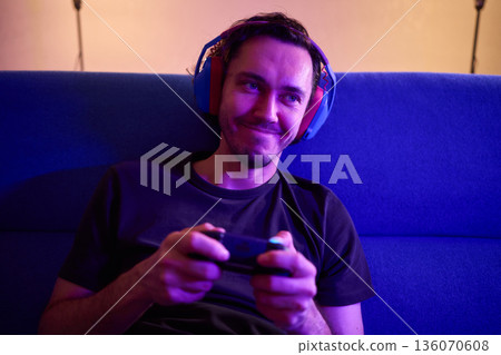 Comfortable man enjoying gameplay under colorful neon illumination, Chill indoor scene featuring smiling individual engaged in video game entertainment amid vivid purple lights 136070608