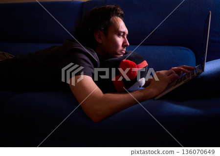 Casual night working from comfy sofa, Quiet evening spent on freelance project in cozy living room, Man enjoying late night work at home with relaxed posture and soft glow 136070649