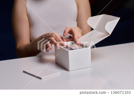 Closeup of hands opening clean packaging under controlled lighting. Lifestyle marketing shot emphasizing tactile clarity while revealing unbranded box in refined setting 136071103