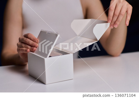 Closeup of hands opening clean packaging under controlled lighting. Lifestyle marketing shot emphasizing tactile clarity while revealing unbranded box in refined setting 136071104