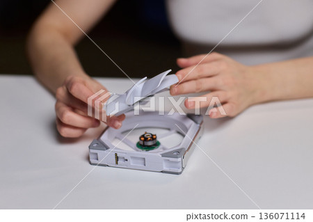 Detailed scene with technician disassembling small desktop fan carefully. Precise hands removing cover of desk fan during technical inspection with focused lighting 136071114
