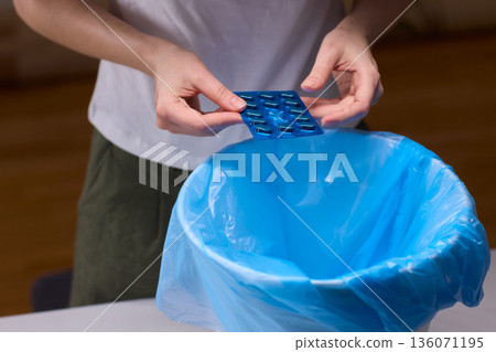 Hands discarding expired pills into bin, closeup of blister pack over blue plastic liner, tabletop setting, focused on safe disposal, mindful household routine, low warm lighting Hands discarding expired pills into bin, closeup of blister pack over blue plastic liner, tabletop setting, focused on safe disposal, mindful household routine, low warm lighting 136071195