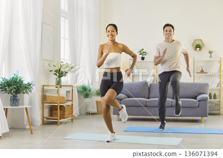 Fit couple doing cardio workout together at home, high knees and smiles indoors Fit couple doing cardio workout together at home, high knees and smiles indoors 136071394