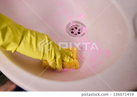 Homeowner prepares tub thoroughly, Closeup of hand cleaning soap scum from bathtub drain, Detailed view of person sanitizing bathtub with pink foam sponge and yellow glove Homeowner prepares tub thoroughly, Closeup of hand cleaning soap scum from bathtub drain, Detailed view of person sanitizing bathtub with pink foam sponge and yellow glove 136071459
