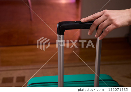 Traveler prepares to leave, Closeup of traveler holding luggage in welcoming hotel entrance, Person grasping suitcase handle in warm hotel lobby signaling upcoming departure 136071585
