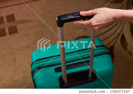 Traveler prepares to leave, Closeup of traveler holding luggage in welcoming hotel entrance, Person grasping suitcase handle in warm hotel lobby signaling upcoming departure 136071587
