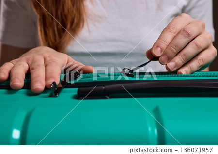 Traveler opening luggage, Close shot of hands unfastening suitcase zipper before travel, Detailed view of hands pulling zipper on vibrant teal travel case with anticipation Traveler opening luggage, Close shot of hands unfastening suitcase zipper before travel, Detailed view of hands pulling zipper on vibrant teal travel case with anticipation 136071957