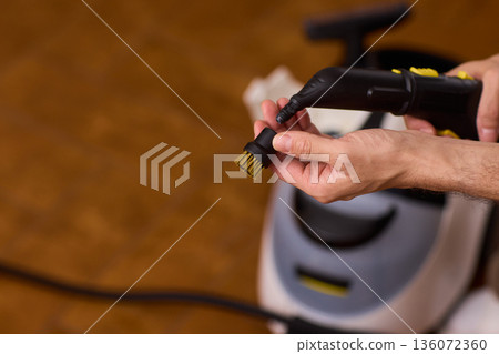 Steam cleaning device being prepared for upholstery and grout cleaning tasks. Focus on textured grip and soft plastic with warm background indicating household maintenance 136072360