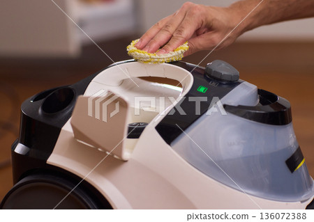 Hand cleaning steam cleaner reservoir with yellow sponge, green indicator light glowing, closeup of plastic tank and handle, focus on routine maintenance and hygiene in modern home Hand cleaning steam cleaner reservoir with yellow sponge, green indicator light glowing, closeup of plastic tank and handle, focus on routine maintenance and hygiene in modern home 136072388